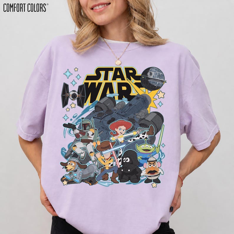 May include: A light purple Comfort Colors t-shirt featuring a Star Wars and Toy Story mashup graphic. The design includes characters like Woody, Buzz Lightyear, and Darth Vader, along with the Star Wars logo and iconic elements.