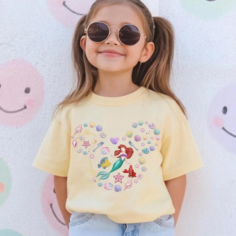 May include: A light yellow t-shirt featuring a heart-shaped design with seashells, starfish, and a mermaid illustration. The design includes a red-haired mermaid, a yellow fish, and a red lobster. The shirt is short-sleeved.