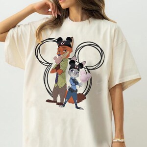 May include: A cream-colored t-shirt featuring a graphic of Nick Wilde and Judy Hopps from Zootopia. Both characters wear black Mickey Mouse ear hats and hold pink Mickey-shaped cotton candy. A large black outline of Mickey ears frames them.
