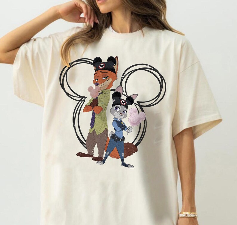 May include: A cream-colored t-shirt featuring a graphic of Nick Wilde and Judy Hopps from Zootopia. Both characters wear black Mickey Mouse ear hats and hold pink Mickey-shaped cotton candy. A large black outline of Mickey ears frames them.