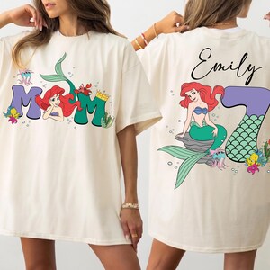 May include: Cream-colored t-shirt featuring a colorful graphic of Ariel from The Little Mermaid. The front of the shirt says "Mom" with Ariel's image. The back of the shirt has Ariel with the number "7" and the name "Emily".