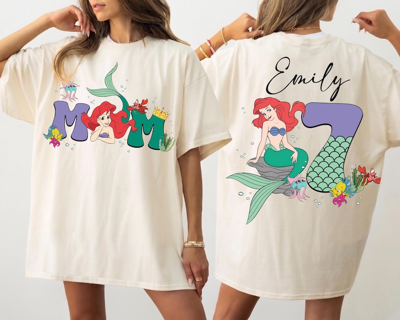 May include: Cream-colored t-shirt featuring a colorful graphic of Ariel from The Little Mermaid. The front of the shirt says "Mom" with Ariel's image. The back of the shirt has Ariel with the number "7" and the name "Emily".