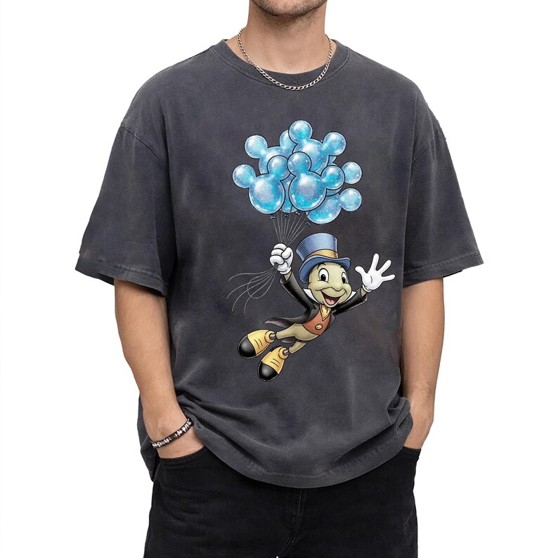 May include: Dark grey t-shirt featuring a cartoon illustration of Jiminy Cricket holding blue Mickey Mouse-shaped balloons. The character is wearing a top hat, suit, and yellow shoes. The shirt has a vintage, washed look.
