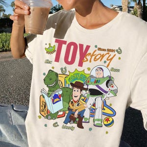 May include: Cream-colored t-shirt featuring a colorful graphic of Toy Story characters, including Rex, Woody, and Buzz Lightyear. The words "Toy Story" are prominently displayed in pink and green, with "Since 2001" above the characters.