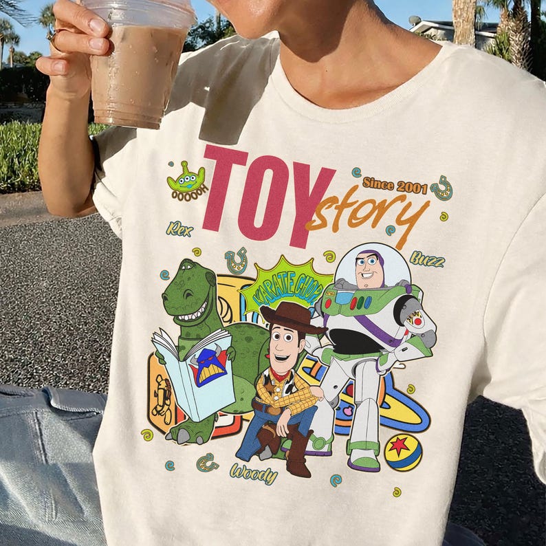 May include: Cream-colored t-shirt featuring a colorful graphic of Toy Story characters, including Rex, Woody, and Buzz Lightyear. The words "Toy Story" are prominently displayed in pink and green, with "Since 2001" above the characters.