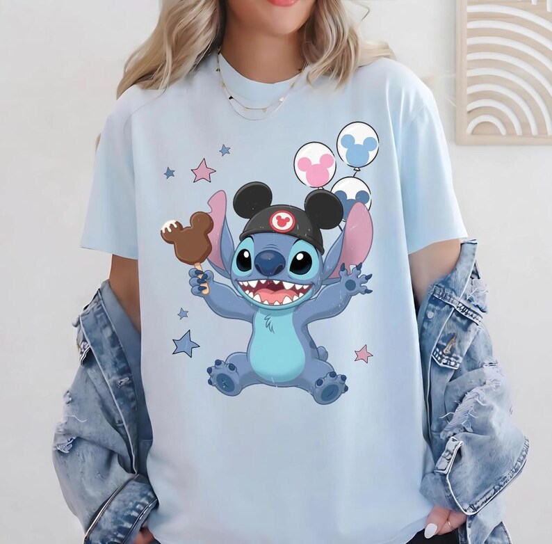 May include: A light blue t-shirt featuring a graphic of the character Stitch wearing a black ear hat with a red icon. Stitch holds a brown mouse-shaped ice cream bar, surrounded by pink and blue stars and three mouse-shaped balloons.