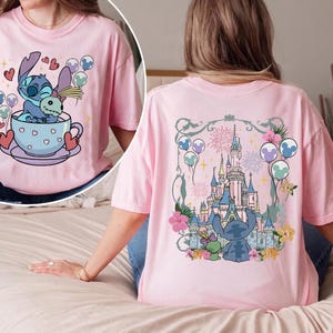 May include: A light pink t-shirt featuring a graphic on the front of a blue alien character sitting in a teacup holding a doll. The back of the shirt displays a large illustration of a castle surrounded by flowers, balloons, and fireworks.