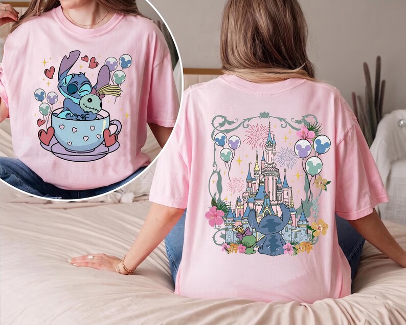 May include: A light pink t-shirt featuring a graphic on the front of a blue alien character sitting in a teacup holding a doll. The back of the shirt displays a large illustration of a castle surrounded by flowers, balloons, and fireworks.