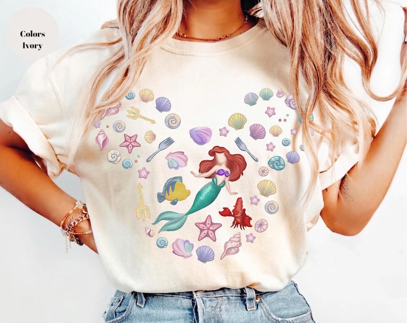 May include: Ivory-colored t-shirt featuring a mermaid design surrounded by seashells, starfish, and other sea-themed illustrations. The mermaid has red hair and a teal tail. The shirt also has the word "Colors" and "Ivory" printed on it.