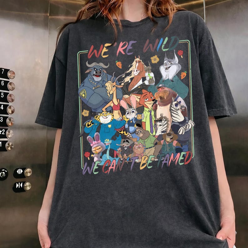 May include: A charcoal gray short-sleeve t-shirt featuring a colorful graphic print of various animated animal characters. The text "WE'RE WILD" is printed at the top and "WE CAN'T BE TAMED" at the bottom in a multi-colored, retro-style font.
