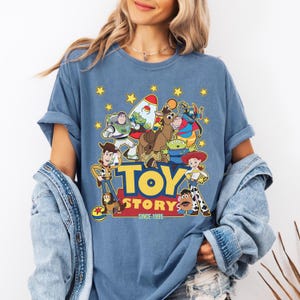 May include: A blue t-shirt featuring a colorful graphic of characters from the animated film "Toy Story." The design includes Woody, Buzz Lightyear, and other characters, with the words "TOY STORY" and "SINCE 1995" in yellow. The shirt has rolled-up sleeves.