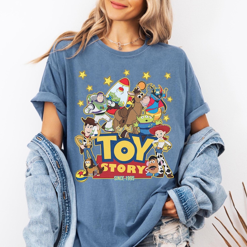 May include: A blue t-shirt featuring a colorful graphic of characters from the animated film "Toy Story." The design includes Woody, Buzz Lightyear, and other characters, with the words "TOY STORY" and "SINCE 1995" in yellow. The shirt has rolled-up sleeves.