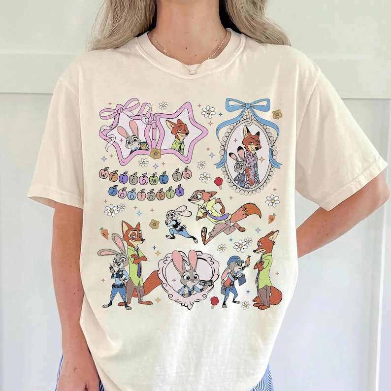 May include: A cream-colored t-shirt featuring a graphic print of characters from Zootopia. The design includes multiple illustrations of a rabbit and a fox, decorative frames, flowers, stars, and the text "Welcome to Zootopia" in colorful letters.
