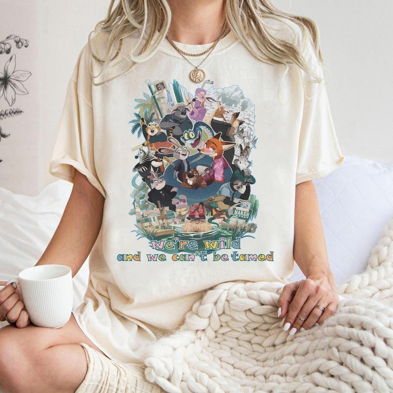 May include: Cream-colored t-shirt featuring a colorful graphic of animated characters. The graphic includes the text "We're wild and we can't be tamed" in a multi-colored font. The shirt has short sleeves and a relaxed fit.