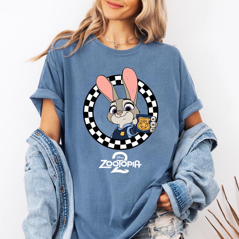May include: A blue t-shirt featuring a graphic of the character Judy Hopps from Zootopia, who is holding a gold police badge inside a black and white checkered circle. Below the character is the white text Zootopia 2 with a Disney logo above it.