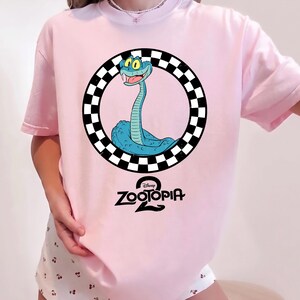 May include: A light pink t-shirt featuring a graphic of a blue snake inside a black and white checkered circle. Below the circle, the text reads Disney Zootopia 2 in a black, stylized font. The shirt has short sleeves and a crew neckline.