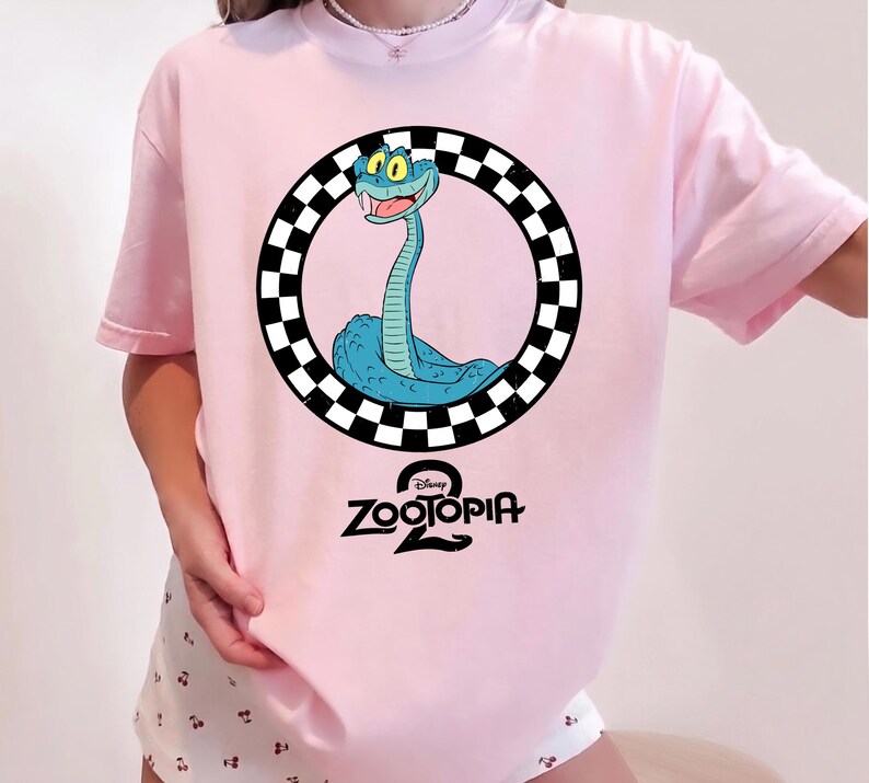 May include: A light pink t-shirt featuring a graphic of a blue snake inside a black and white checkered circle. Below the circle, the text reads Disney Zootopia 2 in a black, stylized font. The shirt has short sleeves and a crew neckline.