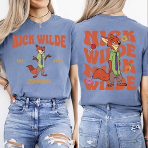 Pode incluir: Uma t-shirt azul com um gr&aacute;fico da personagem Nick Wilde de Zootopia. A frente apresenta o texto NICK WILDE EST. 2016 ZOOTOPIA. As costas mostram um gr&aacute;fico maior de Nick Wilde com o texto repetido NICK WILDE em cor de laranja.