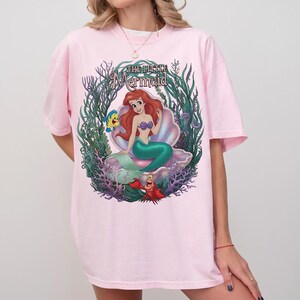 May include: Pink t-shirt featuring a colorful graphic of Ariel from "The Little Mermaid." The design includes Ariel in a seashell, with Flounder and Sebastian. The words "The Little Mermaid" are arched above the image in a script font.