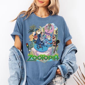 Puede incluir: Camiseta azul con un gr&aacute;fico colorido de la pel&iacute;cula animada "Zootr&oacute;polis". El dise&ntilde;o incluye varios personajes de animales y el t&iacute;tulo de la pel&iacute;cula en verde. La camiseta la lleva una persona con cabello rubio.