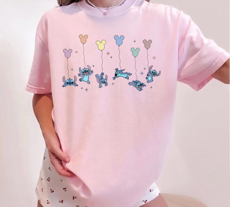 May include: A light pink short-sleeved t-shirt featuring a graphic print of the blue character Stitch from Lilo and Stitch. The character is depicted in various poses, each holding a string attached to a colorful Mickey Mouse-shaped balloon.