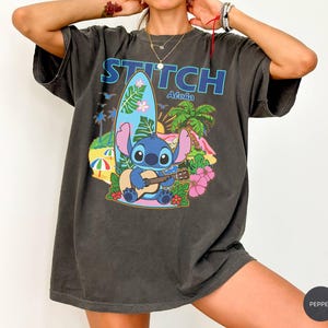 May include: Dark gray t-shirt featuring the word "STITCH Aloha" above a cartoon Stitch character playing a ukulele. The graphic includes a surfboard, palm trees, and flowers, with a beach scene background.