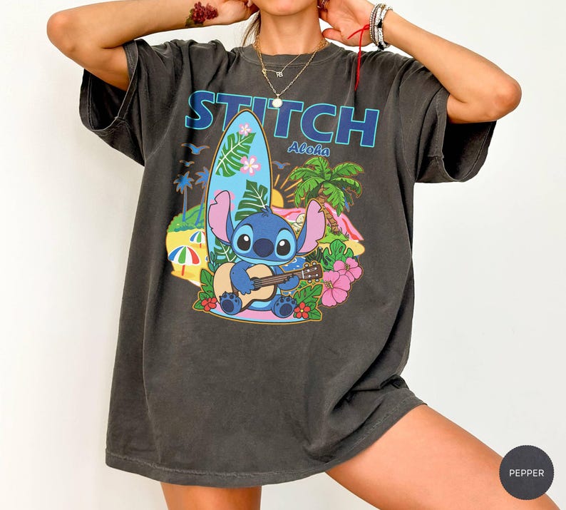 May include: Dark gray t-shirt featuring the word "STITCH Aloha" above a cartoon Stitch character playing a ukulele. The graphic includes a surfboard, palm trees, and flowers, with a beach scene background.