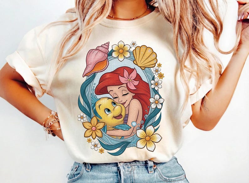 May include: Cream-colored t-shirt featuring a colorful graphic of Ariel from The Little Mermaid hugging Flounder. The design includes seashells, flowers, and a blue oval frame. The shirt is made of a soft material.