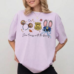 May include: A light purple t-shirt featuring a graphic print of Zootopia-themed icons including a police badge, mouse ears, a carrot, and a blue hat with rabbit ears. The text "Don't mess with the bunny" is printed in black cursive script below.