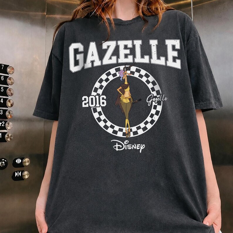 May include: A charcoal grey short-sleeve t-shirt featuring a white graphic print. The design includes the text GAZELLE at the top, the year 2016, a checkered circle, a cartoon gazelle character, the name Gazelle, and the Disney logo at the bottom.