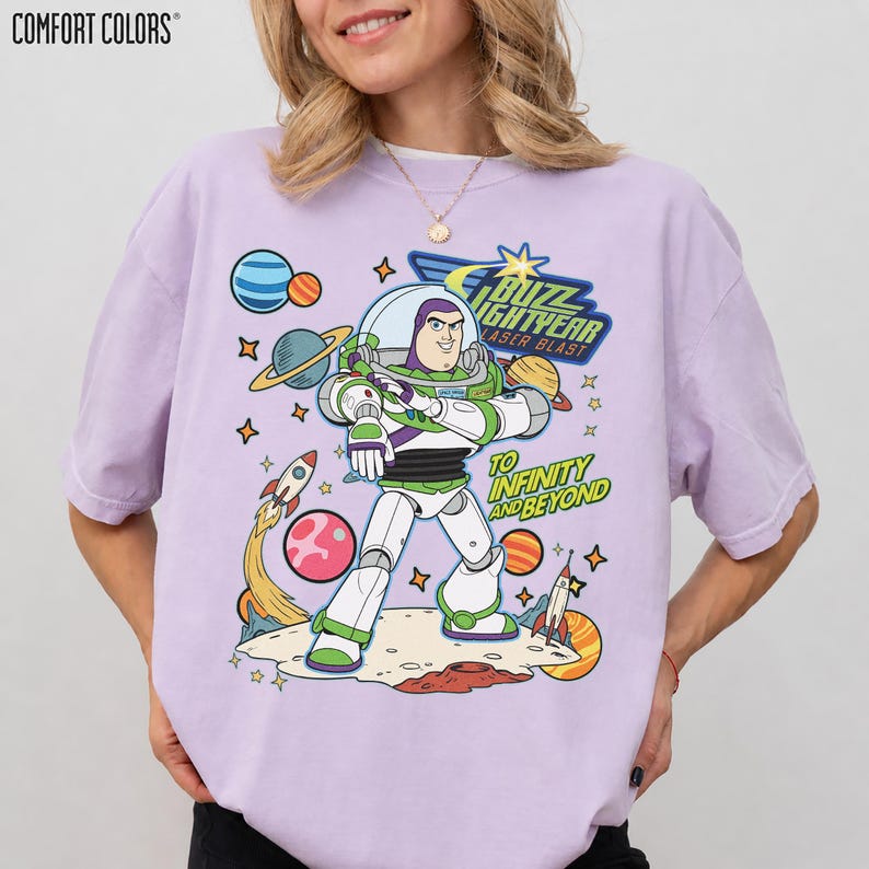 May include: A lilac Comfort Colors t-shirt featuring a colorful graphic of Buzz Lightyear from Toy Story. The design includes the text "Buzz Lightyear Laser Blast" and "To Infinity and Beyond" with planets, rockets, and stars.