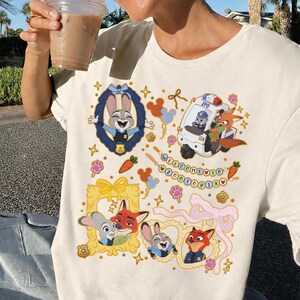 May include: Cream-colored t-shirt featuring colorful cartoon illustrations of characters from Zootopia. The design includes Judy Hopps, Nick Wilde, and a ZPD vehicle, with the text "WELCOME TO ZOOTOPIA".