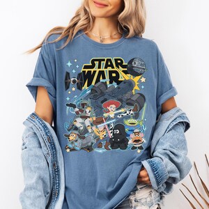 May include: Blue t-shirt featuring a graphic of Star Wars characters and the Star Wars logo. The design includes characters from Toy Story, such as Woody, Buzz Lightyear, and others, combined with Star Wars elements.