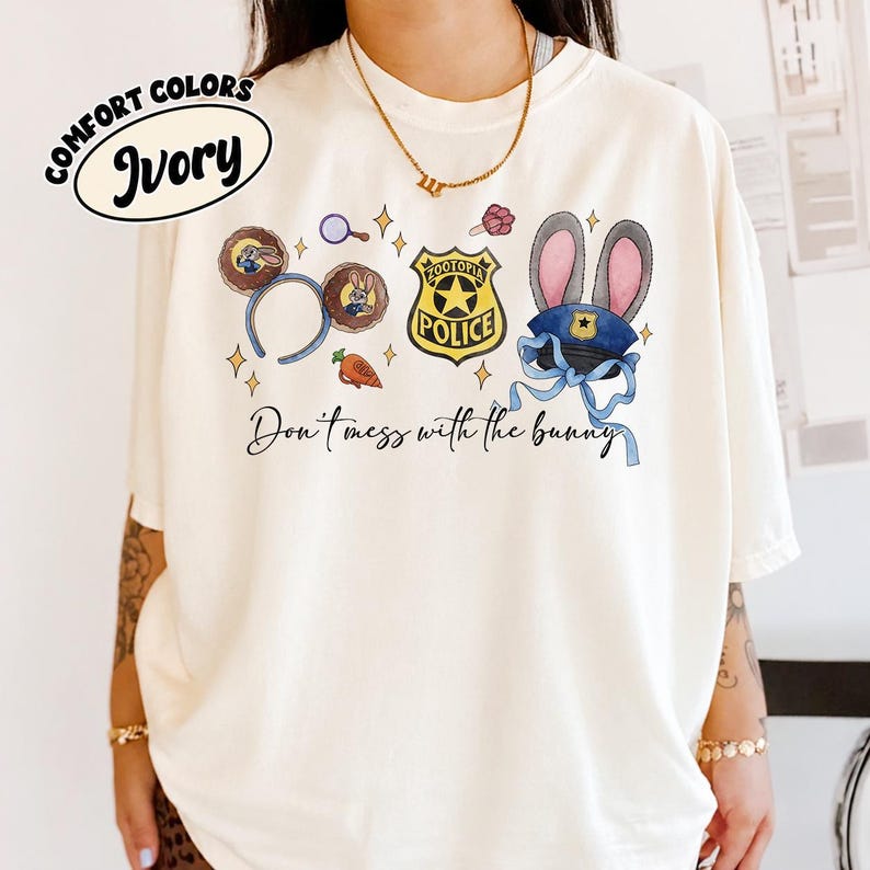 May include: An ivory t-shirt featuring a graphic print with a pair of mouse ears, a Zootopia police badge, a carrot, and a police hat with rabbit ears. The text reads Don't mess with the bunny. The shirt includes a Comfort Colors Ivory label.