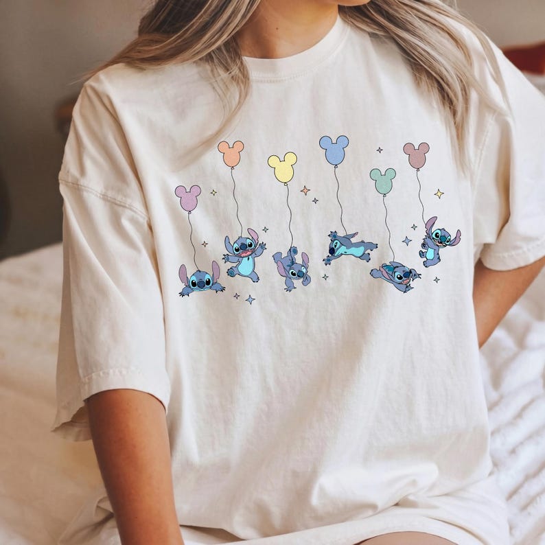 May include: A cream-colored short-sleeve t-shirt featuring a graphic print of the blue character Stitch in various poses. Stitch is shown holding strings attached to colorful Mickey Mouse-shaped balloons in pastel shades, surrounded by small stars.