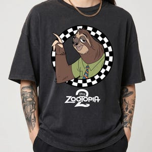 May include: A charcoal gray short-sleeve t-shirt featuring a graphic of the Zootopia sloth character Flash inside a black and white checkered circle. Below the character is the white text Zootopia 2 with the Disney logo above the word Zootopia.