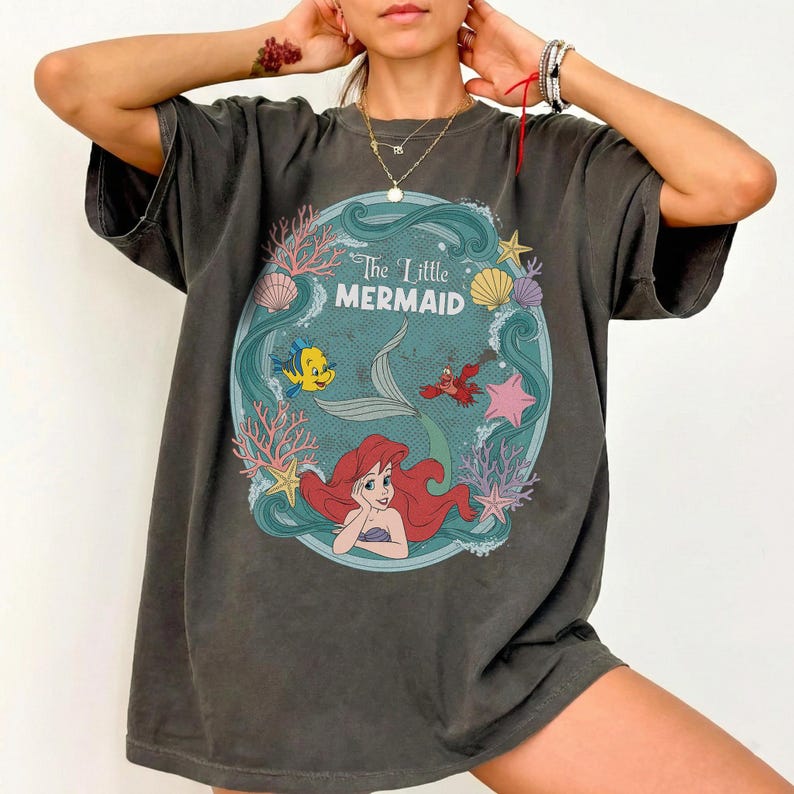May include: Dark gray t-shirt featuring a circular graphic from "The Little Mermaid." The design includes Ariel, Flounder, Sebastian, and other sea elements. The text "The Little Mermaid" is displayed in a teal circle.