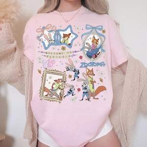 May include: A light pink t-shirt featuring a graphic print of characters from Zootopia, including Judy Hopps and Nick Wilde. The design includes various frames, flowers, and the text Welcome to Zootopia and Zootopia in a colorful, cartoon style.