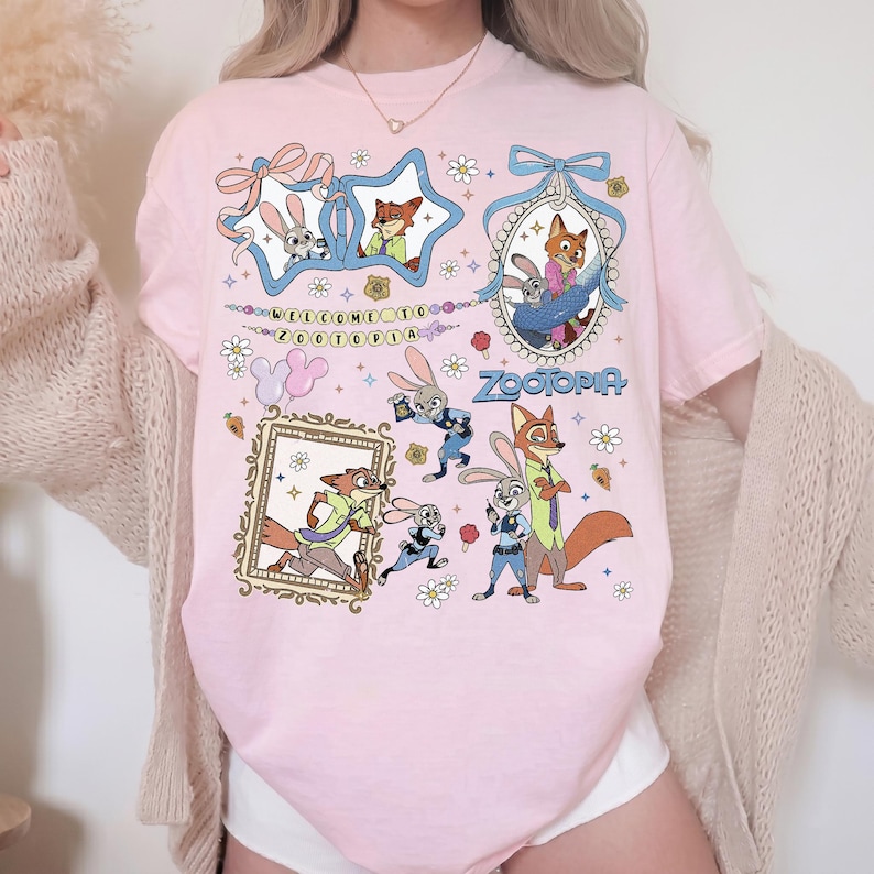 May include: A light pink t-shirt featuring a graphic print of characters from Zootopia, including Judy Hopps and Nick Wilde. The design includes various frames, flowers, and the text Welcome to Zootopia and Zootopia in a colorful, cartoon style.