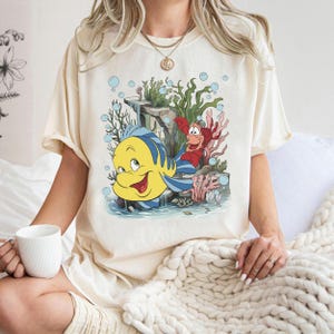 Puede incluir: Camiseta color crema con una ilustraci&oacute;n de dibujos animados de Flounder y Sebasti&aacute;n de La Sirenita. Flounder es amarillo con rayas azules, y Sebasti&aacute;n es rojo. El dise&ntilde;o incluye coral, burbujas y plantas acu&aacute;ticas.