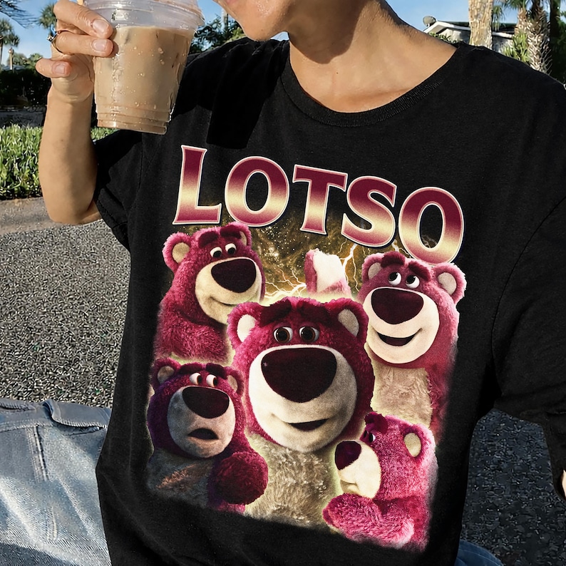 May include: Black t-shirt featuring the word "LOTSO" in a gold and red font above a graphic of several pink teddy bears. The bears have white muzzles and are smiling. The shirt is being worn by a person holding a beverage.