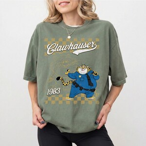 Puede incluir: Una camiseta de manga corta color verde oliva con un gr&aacute;fico de un personaje de guepardo de dibujos animados con uniforme de polic&iacute;a azul. El dise&ntilde;o incluye el texto Clawhauser, el a&ntilde;o 1983 y un patr&oacute;n de cuadros amarillos en el frente.