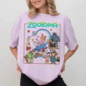 May include: A light purple t-shirt featuring a graphic print of Zootopia characters including a fox, rabbit, sloth, and snake. The design includes the text Zootopia in green letters, surrounded by a thin orange border and small star accents.