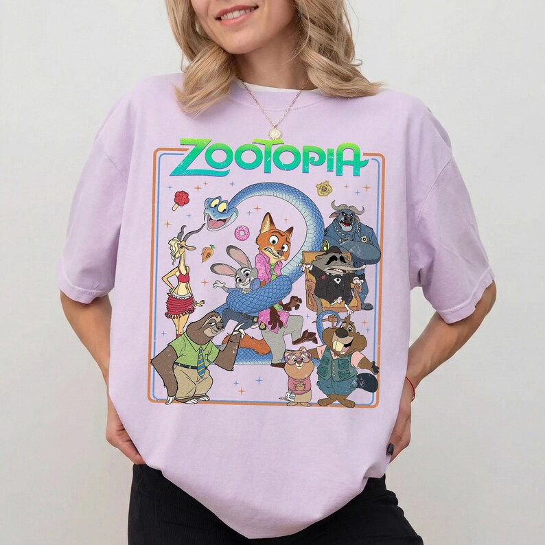 May include: A light purple t-shirt featuring a graphic print of Zootopia characters including a fox, rabbit, sloth, and snake. The design includes the text Zootopia in green letters, surrounded by a thin orange border and small star accents.