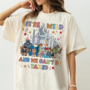 K&ouml;nnte beinhalten: Ein cremefarbenes Kurzarm-T-Shirt mit einer bunten Grafik verschiedener animierter Tierfiguren vor einer Stadtsilhouette. Der Text "WE'RE WILD AND WE CAN'T BE TAMED" ist in mehrfarbigen Buchstaben &uuml;ber und unter den Charakteren aufgedruckt.
