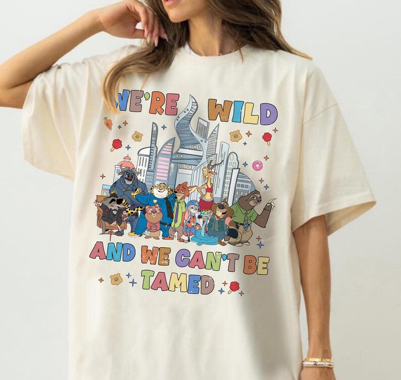 K&ouml;nnte beinhalten: Ein cremefarbenes Kurzarm-T-Shirt mit einer bunten Grafik verschiedener animierter Tierfiguren vor einer Stadtsilhouette. Der Text "WE'RE WILD AND WE CAN'T BE TAMED" ist in mehrfarbigen Buchstaben &uuml;ber und unter den Charakteren aufgedruckt.