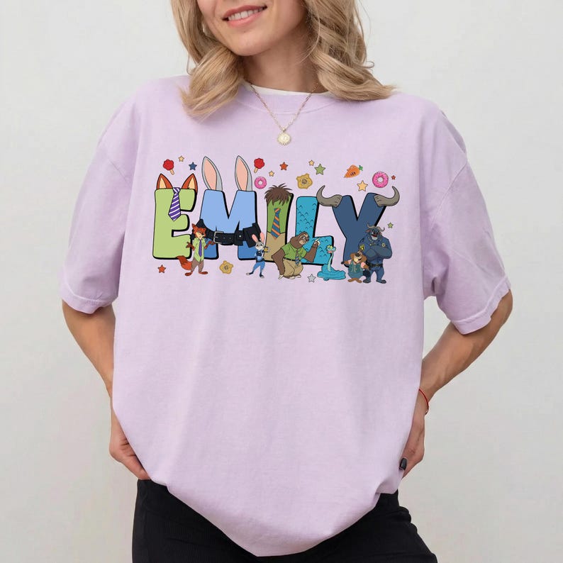 Pode incluir: Uma t-shirt de manga curta em roxo claro com o nome EMILY em letras grandes e coloridas. Cada letra incorpora personagens animais animados e acess&oacute;rios, incluindo orelhas, gravatas e pequenos &iacute;cones como estrelas e donuts.