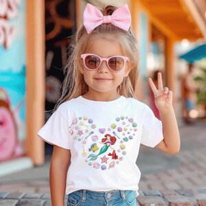 May include: A young girl wearing a white t-shirt with a colorful mermaid design, pink sunglasses, and a large pink bow. The t-shirt features a heart-shaped design with seashells, a mermaid, and other sea-themed elements.