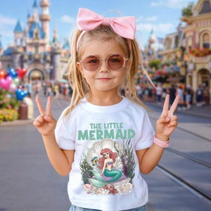 May include: A young girl wearing a white t-shirt with the words "THE LITTLE MERMAID" in green. The shirt features a graphic of Ariel in a clam shell. She wears a pink bow, sunglasses, and a pink bracelet. She is giving the peace sign.