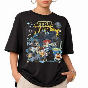May include: Black t-shirt featuring a graphic of Star Wars characters and the text "STAR WARS" in yellow. The graphic includes characters from Toy Story and Star Wars, with a spaceship and stars.